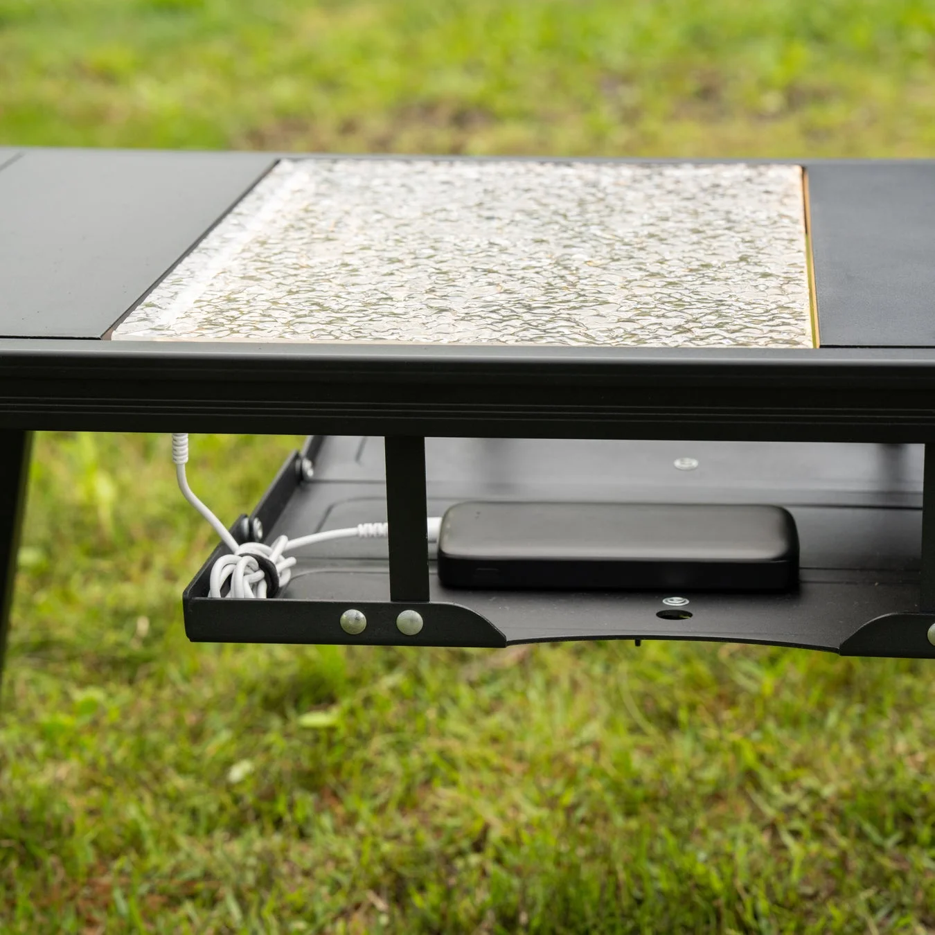 屋外用の黒いテーブルの下にモバイルバッテリーと充電ケーブルが収納されている様子。テーブルの天板は質感のあるガラス製で、背景には緑の芝生が広がっています。