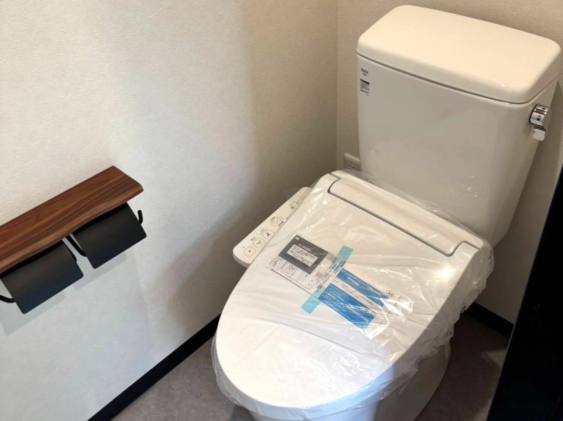 新しいかリフォームされたばかりの清潔なトイレ空間です。保護ビニールに覆われたINAX製のウォシュレット付き便器と、木製棚付きのスタイリッシュな2連トイレットペーパーホルダーが設置されています。