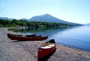 晴れた日の湖畔に赤いカヌーが2艘置かれ、遠くには緑豊かな山々が広がる、穏やかで美しい自然の風景です。
