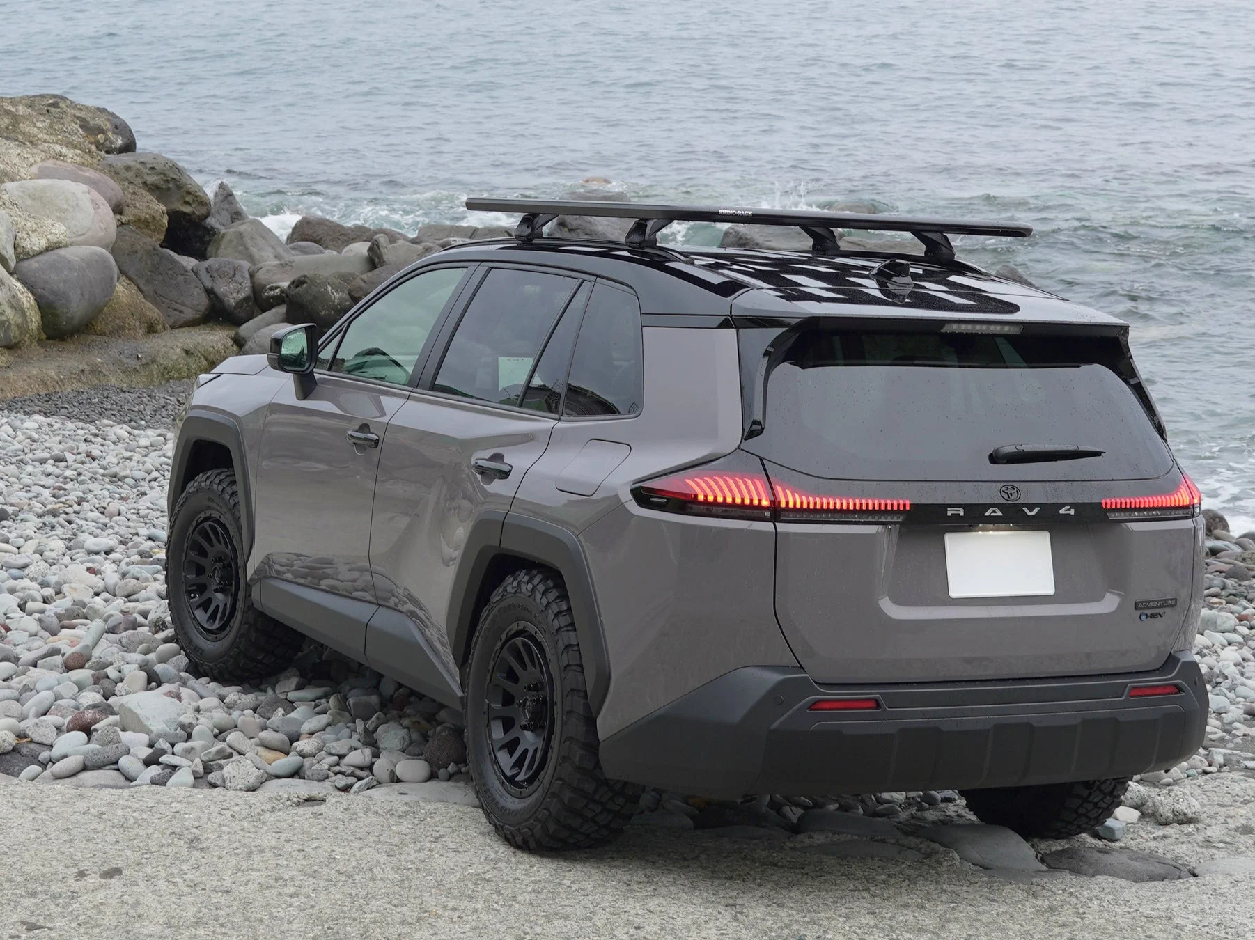 グレーのトヨタRAV4が、岩と小石の多い海岸に駐車されています。車両にはルーフラックが装備され、背景には波打つ海が広がっています。