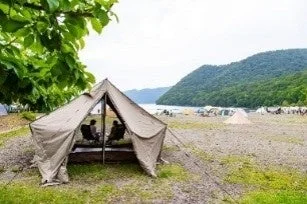 湖畔のキャンプ場で、大きなテントの中に座る二人の人物が見えます。周囲には他のテントも点在し、山々に囲まれた自然豊かな風景が広がっています。
