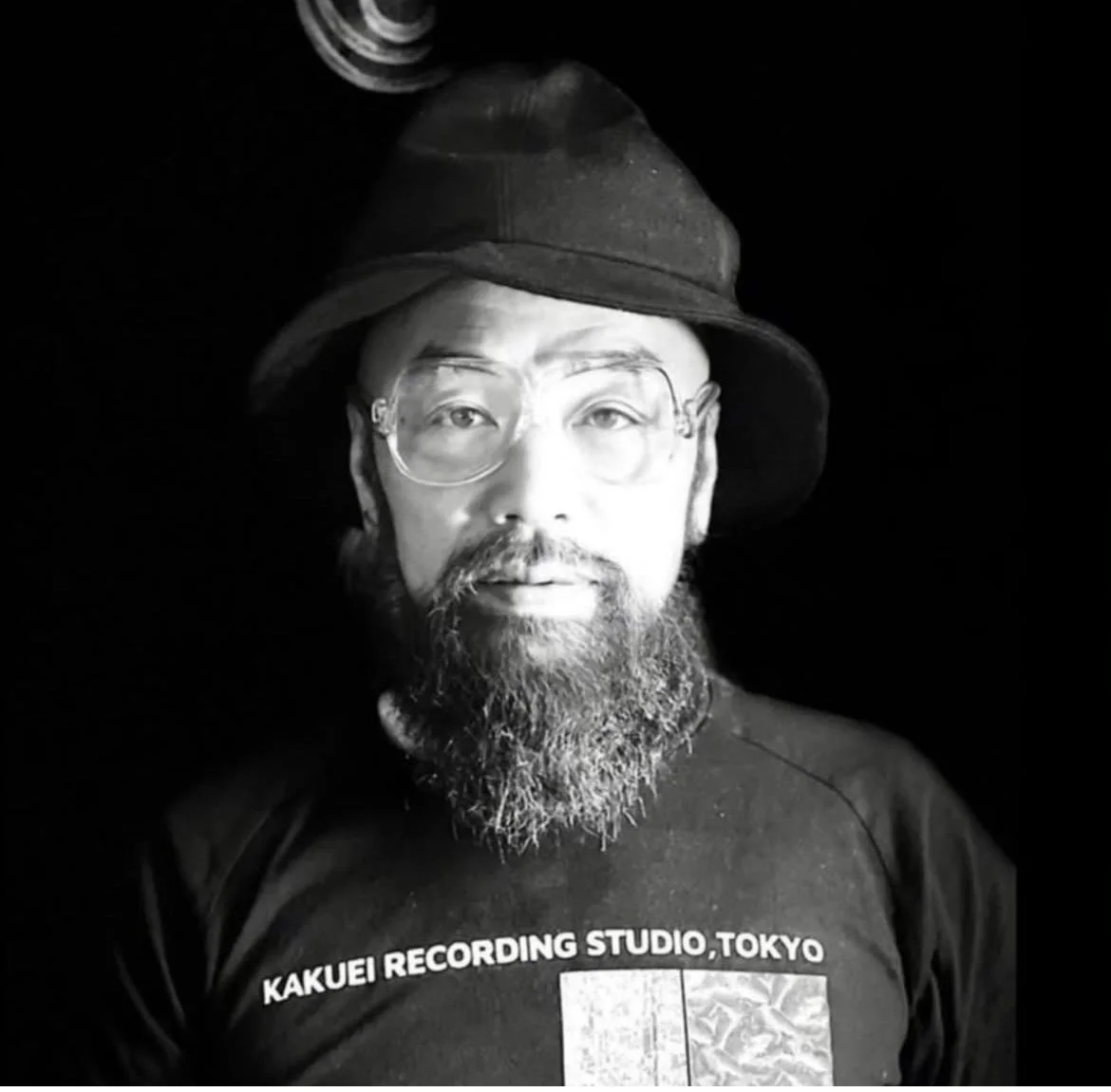 黒い帽子と眼鏡をかけたひげの男性が正面を向いているモノクロ写真。Tシャツには「KAKUEI RECORDING STUDIO, TOKYO」と書かれている。