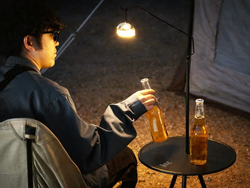 夜のキャンプ場で、男性が椅子に座り、ランタンの光の下でビールを飲んでいる様子