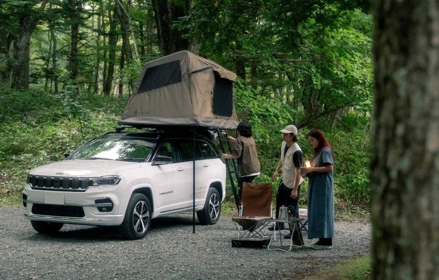森林の中で白いJeep Commanderにルーフトップテントを設置し、人々がキャンプを楽しんでいる様子