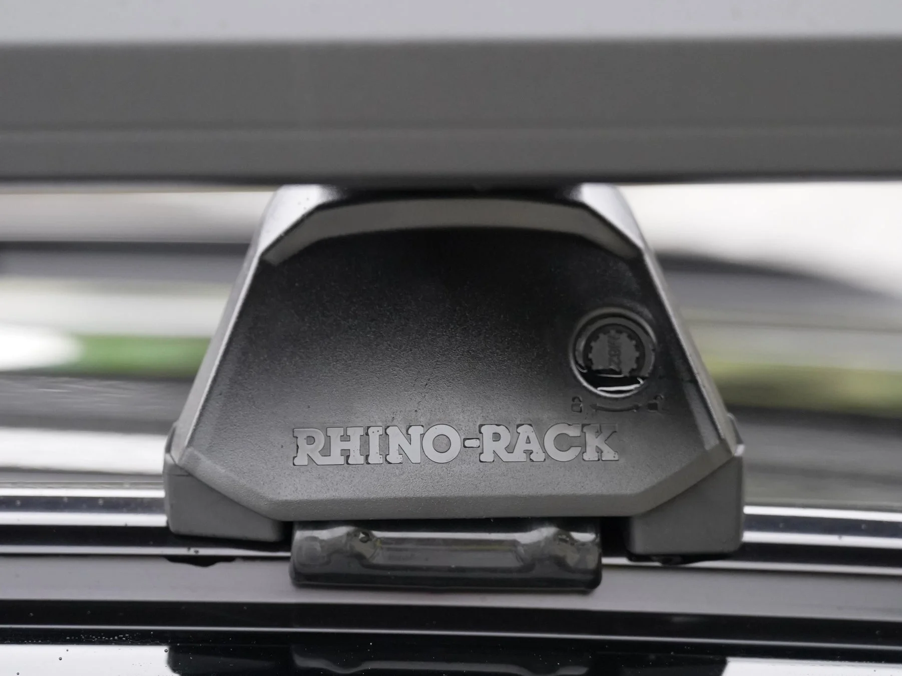 車のルーフラックのクローズアップ画像です。ルーフラックの部品には「RHINO-RACK」というブランド名が刻印されており、ロック機構のような丸い部分も見えます。