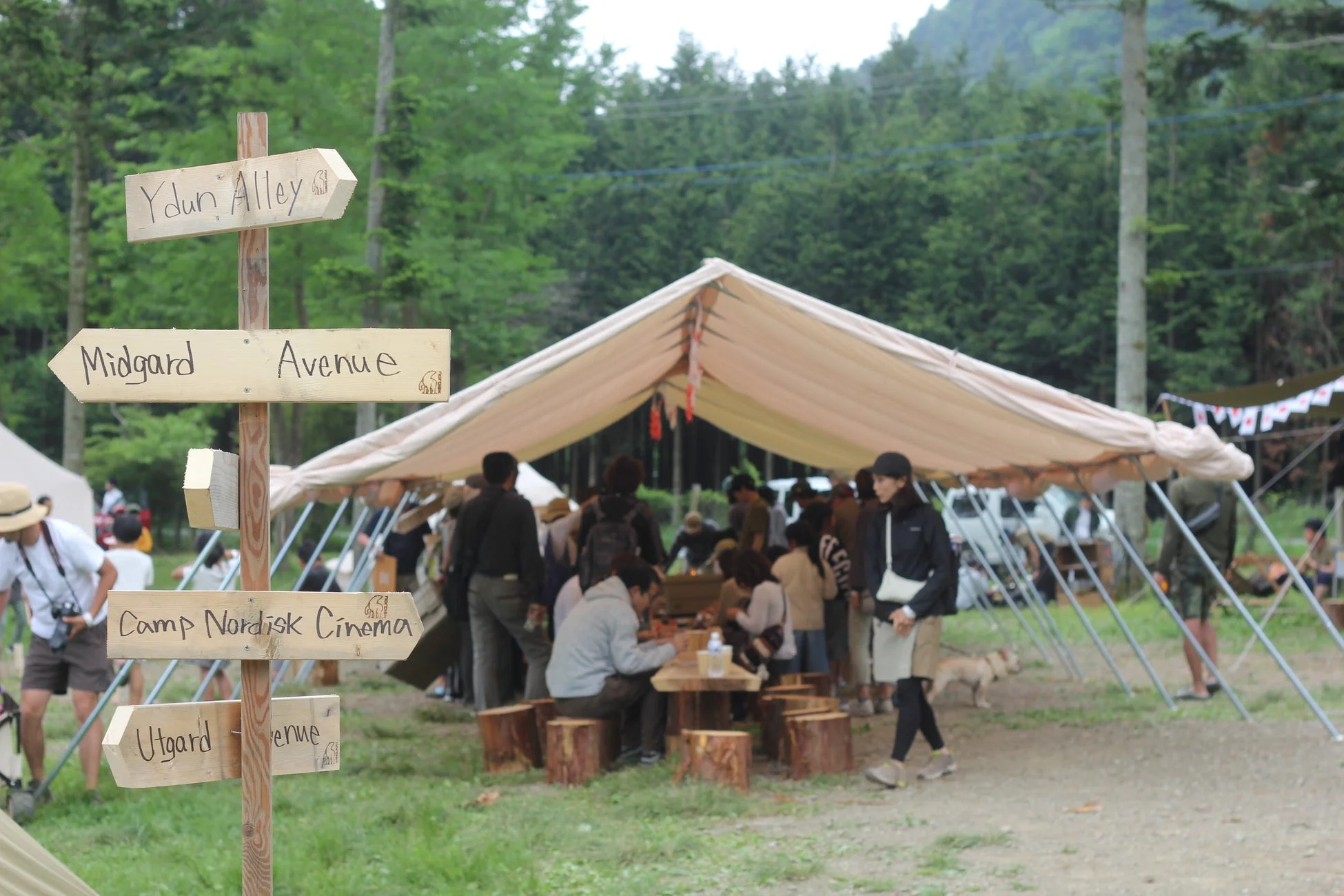 屋外イベントの様子を捉えた画像。木製の案内板には「Ydun Alley」「Midgard Avenue」「Camp Nordisk Cinema」と表示され、大きなテントの下には人々が集い、丸太のテーブルと椅子でくつろいでいる。自然豊かな森の中で、多くの人が楽しんでいる様子がうかがえる。