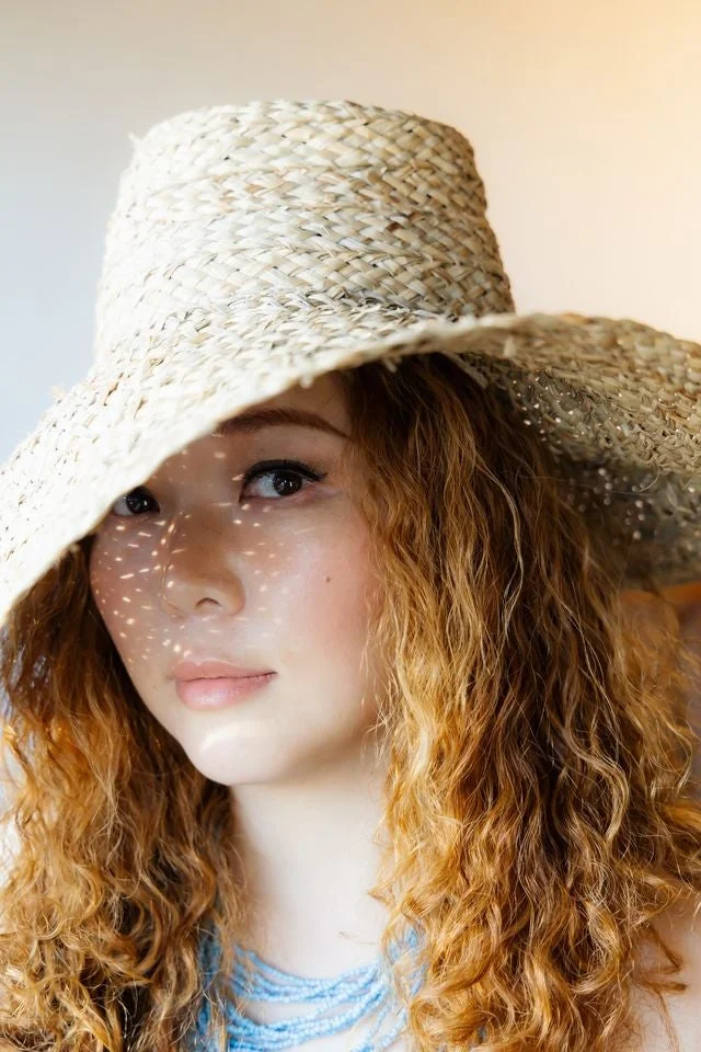 麦わら帽子をかぶり、日差しが顔に影のパターンを作る女性のポートレート。ウェーブのかかった髪とビーズのネックレスが特徴的で、自然な美しさを引き立てています。