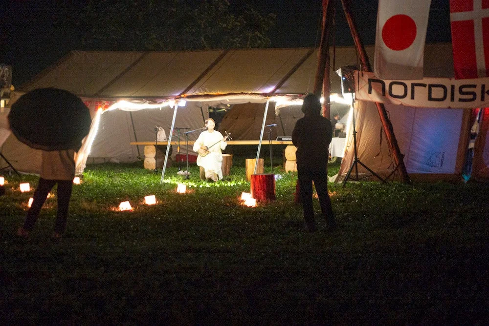 夜の野外イベントで、テントの前で民族楽器を演奏する人物と、それを見守る人々が写っています。足元にはランタンが灯り、日本国旗も掲げられています。