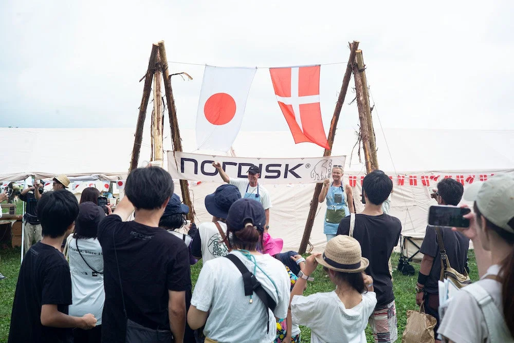 屋外で開催されているアウトドアイベントの様子。日本の国旗とデンマークの国旗、NORDISKのバナーが掲げられ、多くの人々が集まっている。