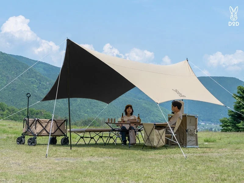 晴れた日に山を背景にした広々とした芝生で、DODのタープの下でカップルがリラックスしてキャンプを楽しんでいる様子