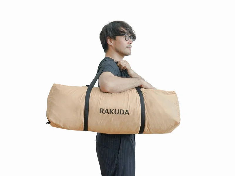 メガネをかけた男性が、肩に「RAKUDA」と書かれたベージュ色の大きな収納バッグを担いでいる