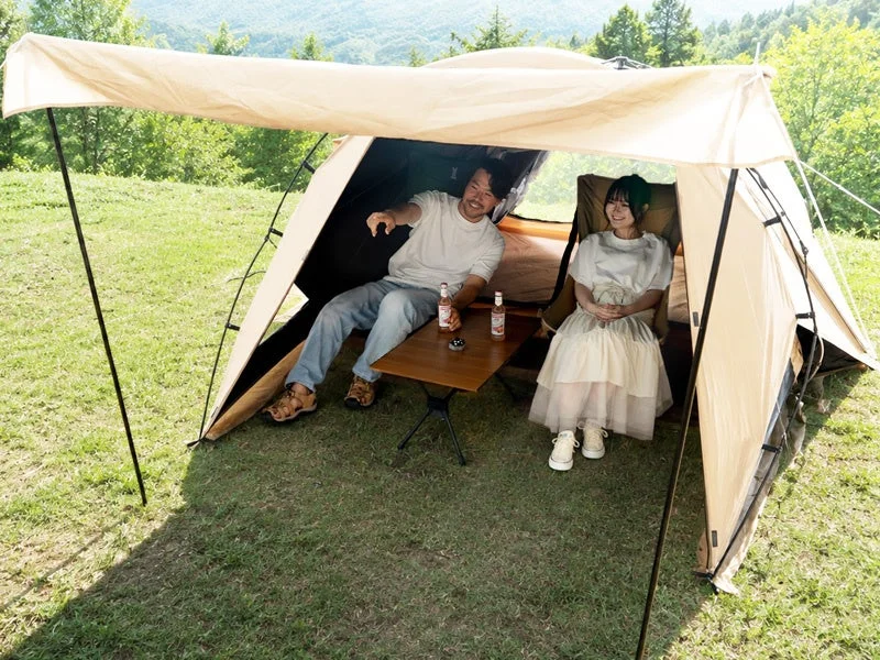 自然豊かな山と森を背景に、ベージュのテントの下で男女がリラックス
