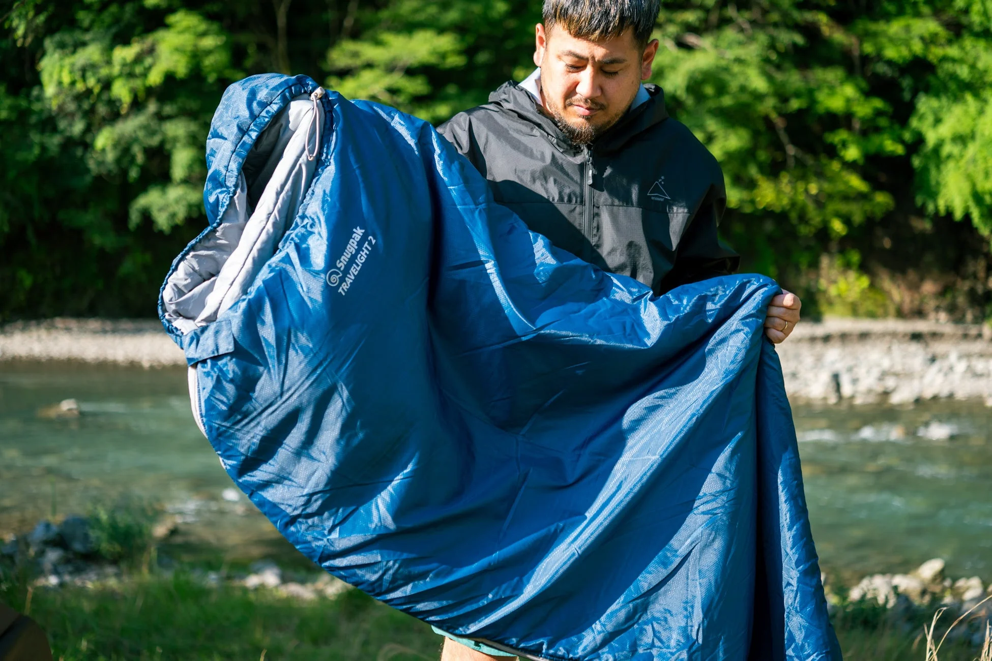 男性が川辺の自然の中で、青い寝袋「Snugpak TRAVELIGHT 2」を広げている様子